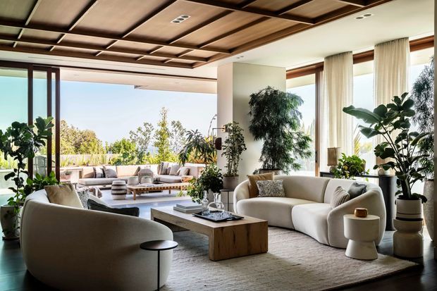 A bright, spacious contemporary living room with extensive floor-to-ceiling glazing opening toward a landscaped terrace. The room features sculptural cream sofas and armchairs arranged around a low timber coffee table, abundant indoor plants, sheer curtains, and a dramatic grid-pattern timber ceiling with recessed lighting. The neutral palette and natural light create a refined, inviting atmosphere.