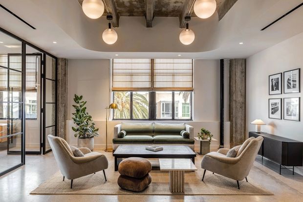 A refined contemporary living room with a neutral palette, two upholstered lounge chairs, a low green sofa, layered coffee tables, and a dark sideboard. Black-framed glazed partitions sit to the left, while the ceiling features exposed concrete or timber-like structural beams and spherical pendant lights. Full-height windows with woven blinds bring in natural light, and framed artwork decorates the right wall.