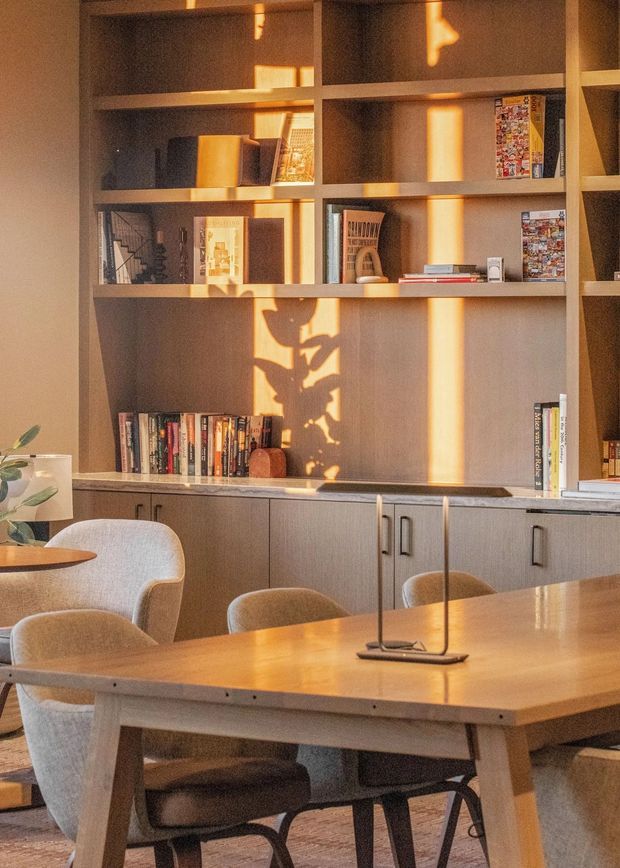 Warmly lit interior featuring a timber dining table with upholstered dining chairs in front of a full-height built-in shelving and cabinetry wall. The shelving displays books and small decorative objects, while low-angle sunlight creates strong geometric shadows across the joinery. The space feels like a refined dining and study area with muted timber, beige, and warm neutral tones.