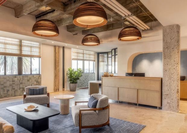 A warm, contemporary reception or lounge interior with exposed concrete structure, industrial ceiling services, large windows, a timber reception counter, and several sculptural pendant lights. The space includes upholstered lounge chairs, a low coffee table, a patterned rug, indoor planting, and textured tiled wall sections. Rounded architectural openings and layered natural materials create a refined hospitality or commercial atmosphere.