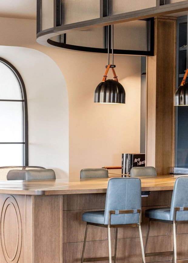 A refined dining or entertaining area with a long timber island or bar-height counter, integrated wood cabinetry, upholstered blue-grey stools, and a ribbed dark metal pendant light suspended by orange leather straps. The room features a large arched window with black framing, warm beige walls, and a curved overhead architectural screen or partition with fluted translucent panels. The overall composition is warm, tailored, and contemporary with subtle Art Deco-inspired curves and detailing.