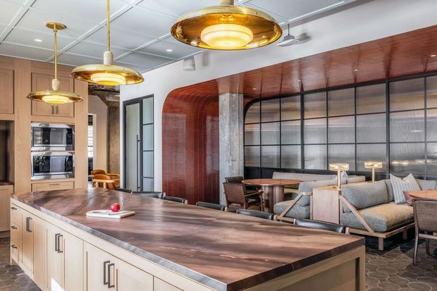 A sophisticated open-plan kitchen and living area with a large dark timber island, light built-in cabinetry, integrated appliances, and distinctive brass pendant lights. The adjacent lounge features timber-framed seating, patterned upholstery, and small table lamps. A curved red-brown feature wall and ceiling treatment frames a dark metal and glass partition, while exposed concrete columns and geometric flooring add an industrial architectural character.
