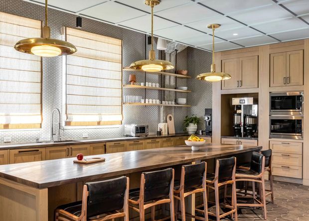 A spacious, warm-toned kitchen with a large dark-stained island and seating for several people. The room features extensive natural timber cabinetry, integrated stainless-steel appliances, open timber shelving, patterned tile splashbacks, woven window shades, and prominent brass pendant lights. The kitchen has a refined contemporary look with traditional timber detailing and a welcoming entertaining layout.