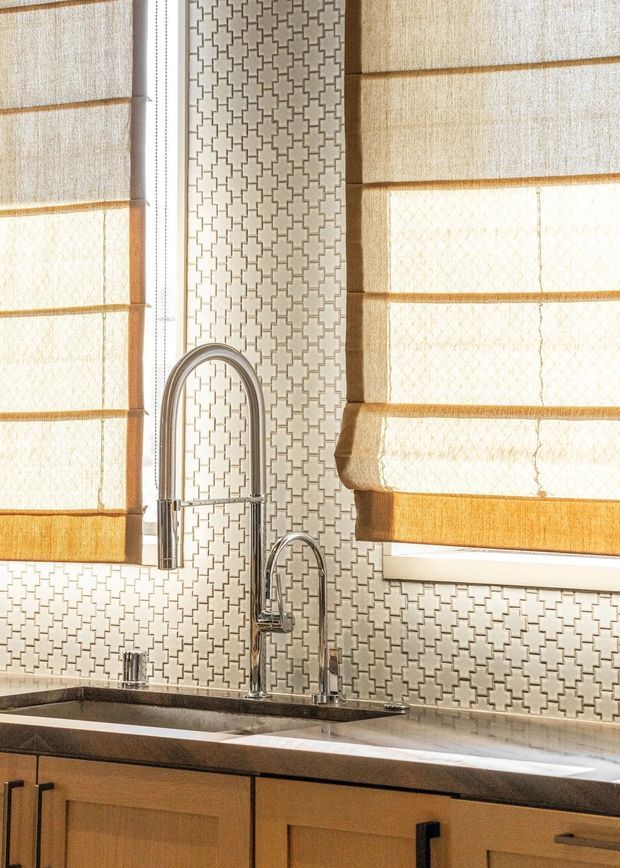 Close-up of a warmly lit kitchen sink area with two chrome gooseneck faucets, a dark countertop, natural timber cabinetry, and a repeating geometric patterned tile backsplash. Cream Roman shades cover the surrounding windows, creating a bright, refined traditional interior.