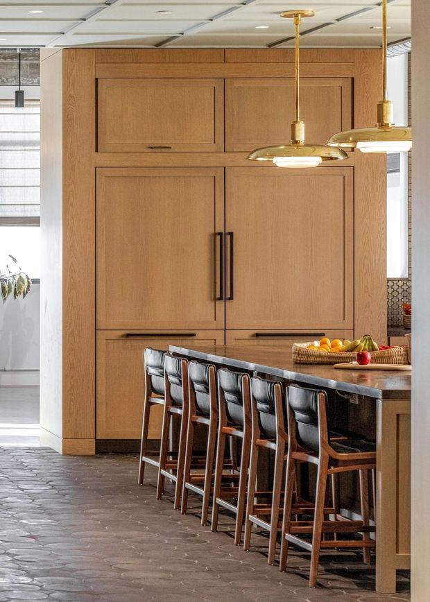 A warm, refined kitchen interior featuring a full-height light timber built-in wall with integrated cabinet and appliance doors, dark stone island benchtop, upholstered timber bar stools, and two brass pendant lights. The floor has dark irregular stone or slate-like paving, while large windows bring in natural light. The image is composed primarily around the kitchen island and cabinetry.