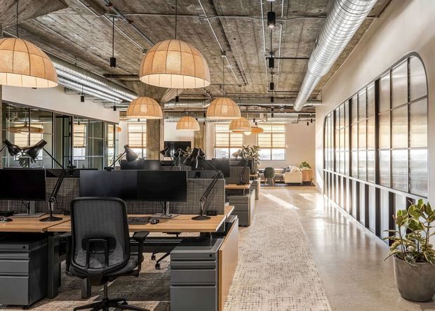 A bright open-plan commercial office with rows of workstations, computer monitors, ergonomic chairs, and desks. The space features exposed concrete ceilings, visible ductwork, black-framed industrial-style glazing, pendant lights with woven dome shades, and a broad central circulation area. Neutral finishes and abundant daylight create a contemporary industrial workplace atmosphere.