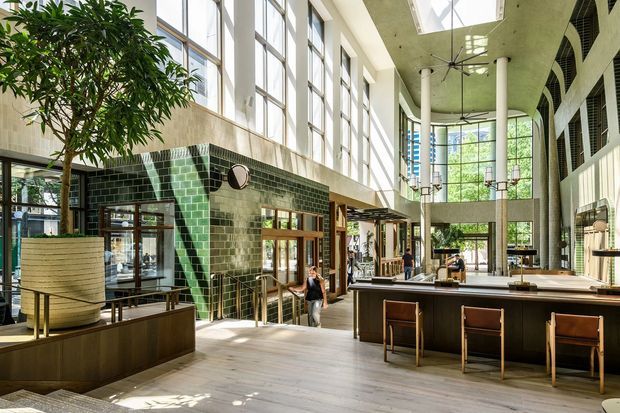 A bright, expansive commercial interior resembling a hotel or office lobby, with a double-height glazed atrium, tall clerestory windows, skylights, exposed white columns, and a large central bar or reception counter. Dark green glazed brick forms a prominent tiled wall, while timber-framed doors, dark built-in joinery, pale flooring, and indoor planting contribute to the contemporary architectural character.