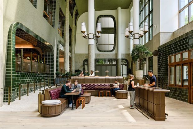 A tall, double-height hospitality interior with a refined Art Deco-inspired design. The space features dark green glazed tilework wrapping the walls and arched openings, pale timber flooring, large steel-framed windows, decorative chandeliers, a central bar or reception counter, and burgundy upholstered banquette seating. People are gathered informally around lounge and dining tables, creating an inviting hotel-lobby or upscale café atmosphere.