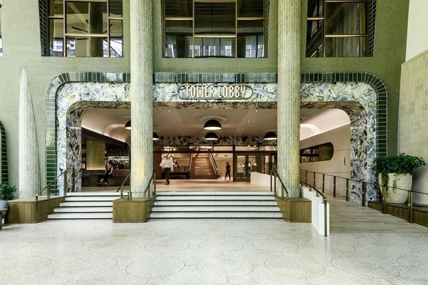 Grand commercial tower lobby entrance with broad terrazzo steps, tall cylindrical mosaic-clad columns, green textured wall surfaces, ornate marble-patterned portal detailing, and a visible reception or circulation area beyond. The space has a strong mid-century-inspired commercial interior character with dramatic scale and layered architectural detailing.