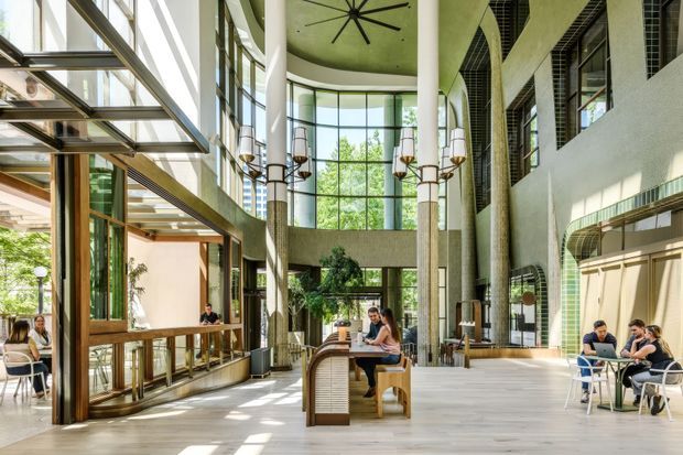 A spacious, sunlit multi-level commercial interior with a tall atrium, floor-to-ceiling glazing, and a central communal timber worktable. The space includes café-style seating, integrated counters, cylindrical columns, dramatic pendant lights, a large ceiling fan, and a curved feature ceiling. Exposed structural and mechanical elements, pale timber flooring, timber-framed glazing, and greenish tiled wall surfaces create a contemporary hospitality or workplace environment.