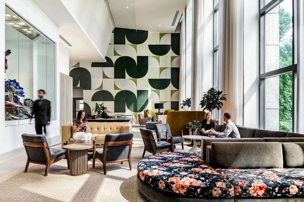 A bright, spacious contemporary lounge or hotel-style lobby with floor-to-ceiling black-framed glazing, pale flooring, and varied seating arranged around low tables. A large circular upholstered sofa with a bold floral pattern dominates the foreground, while the rear wall features a striking oversized geometric mural in dark green, black, cream, and grey. Indoor plants, artwork, and layered lounge seating create an inviting social space.