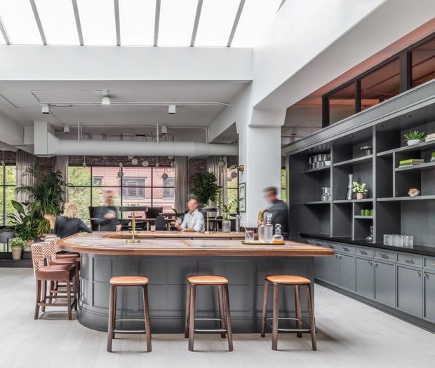 A bright, open-plan commercial hospitality space centred on a large dark grey bar island with a timber countertop and three stools. Built-in shelving and cabinetry line the right wall, displaying glassware and plants, while additional seating, pendant lighting, exposed services, greenery, and large industrial-style windows create a contemporary café or restaurant atmosphere.