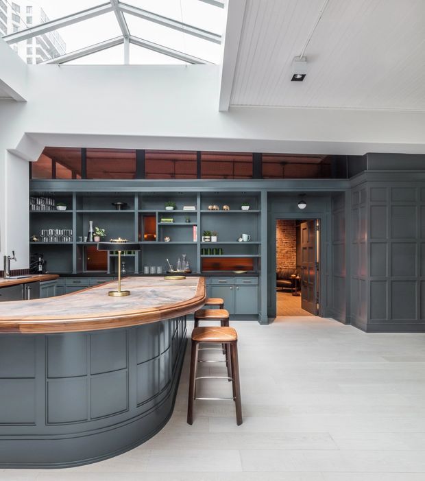 A sophisticated, spacious kitchen with a large curved island and integrated bar seating. The room features dark blue-grey panelled cabinetry, open shelving, warm timber accents, and a pale stone benchtop. A glazed roof lantern brings abundant natural light into the space, while a doorway leads to a warmly lit adjoining room. The design combines refined traditional joinery with contemporary industrial-style metal framing and warm wood details.