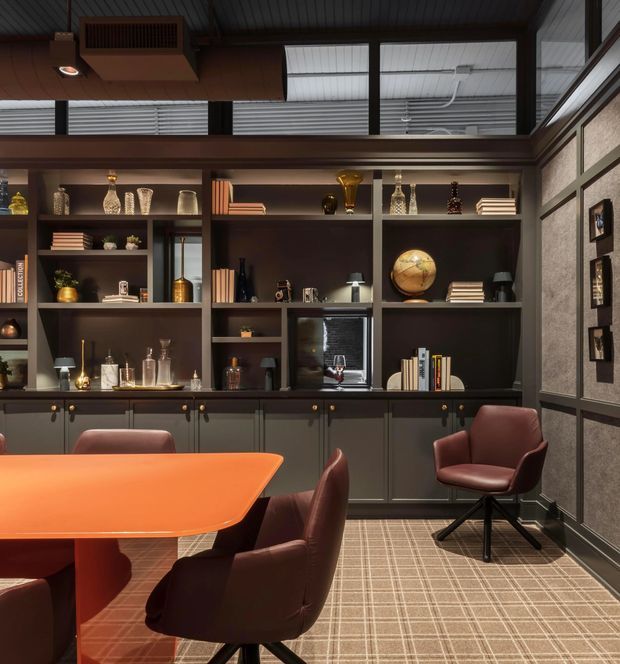 A richly styled study or library with dark built-in shelving and lower cabinetry spanning the back wall, displaying books and decorative objects. A large orange table with burgundy upholstered chairs occupies the foreground. The room features a cofferless dark ceiling with clerestory glazing, framed textured wall panels, integrated shelf lighting, and patterned carpet, creating a moody, sophisticated workspace.