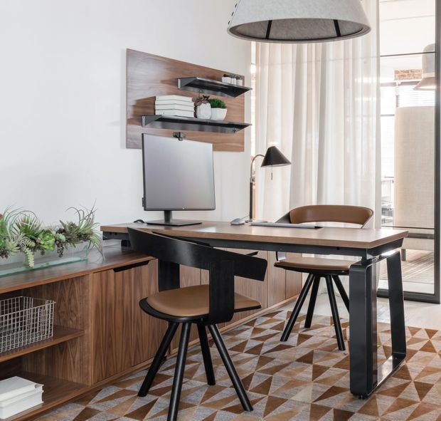 A stylish home office with a dark timber desk, computer monitor, two brown leather or faux-leather chairs, and a wall-mounted shelving system set against a timber feature panel. A low timber sideboard with open shelving occupies the left side, while sheer curtains and a glass door or window provide abundant natural light. The room has a patterned geometric area rug and a large drum pendant light, creating a refined mid-century-inspired workspace.