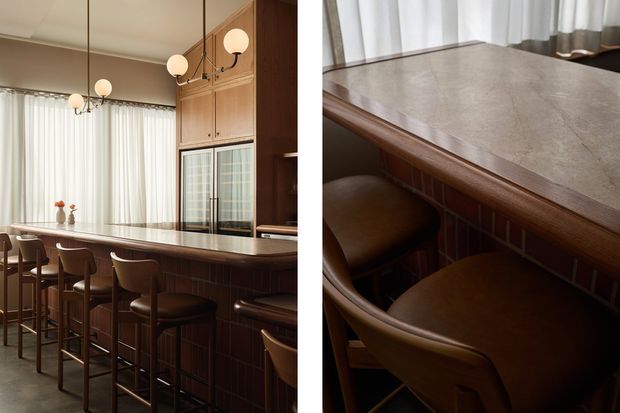 A warm, refined kitchen or dining space centred on a long island or breakfast bar with a light stone benchtop, dark timber cabinetry, and integrated appliances. Several brown upholstered bar stools line the island beneath globe pendant lights. Floor-to-ceiling sheer curtains provide bright natural light, while the close-up view highlights the stone surface, timber edge detailing, and curved stools.
