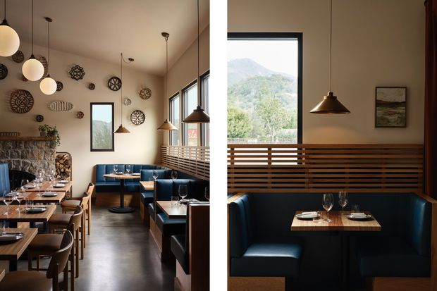 A warmly lit contemporary restaurant interior with blue upholstered booth seating, timber tables, pendant lights, polished concrete flooring, and a stone-clad feature wall. Decorative circular wall pieces and framed artwork add character. Large windows provide views toward a green mountainous landscape, while timber horizontal screening separates dining areas.