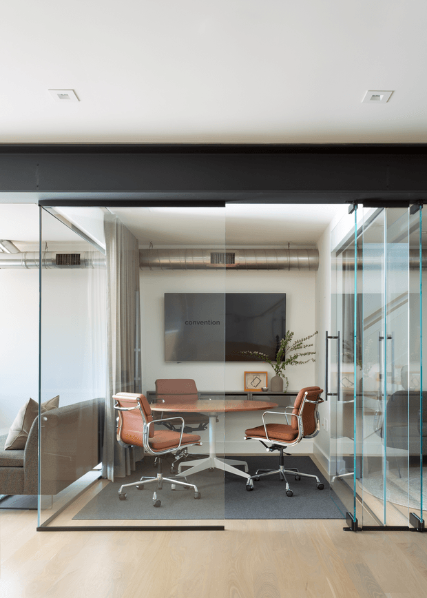 A compact glass-enclosed office or meeting room with transparent sliding glass doors, a round wood-toned meeting table, four leather office chairs, and a wall-mounted display. The room has exposed silver ductwork, dark structural beams, pale timber flooring, neutral walls, and a soft contemporary workplace aesthetic. A lounge area is visible beyond the glazing.
