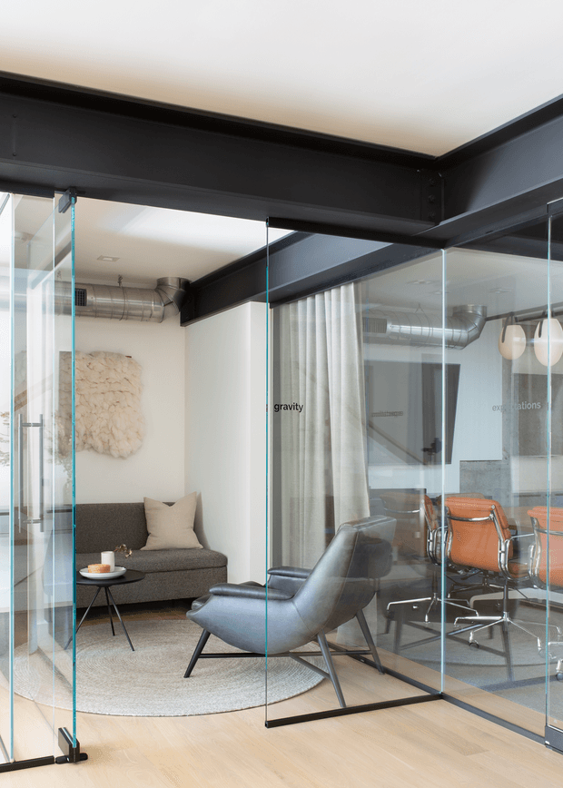 An open-plan contemporary office interior with a glass-enclosed meeting room and a small lounge area. Exposed dark steel structural beams and exposed silver ventilation ducts contrast with white walls and a pale timber floor. The lounge includes a dark grey sofa, black leather lounge chair, round coffee table and light neutral rug, while the meeting room contains a table and orange leather office chairs. Full-height clear glass partitions and doors provide visual connection between the spaces.