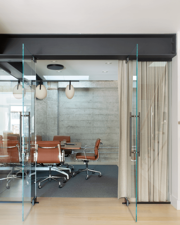 Interior view of a modern glass-enclosed meeting room or private office. The space features a raw exposed concrete feature wall, dark ceiling bulkhead, full-height clear glass doors and partitions, beige curtains, warm timber flooring, dark carpet, and a set of brown leather office chairs around a conference table. Minimal pendant lights and recessed ceiling lighting create a refined contemporary workplace setting.