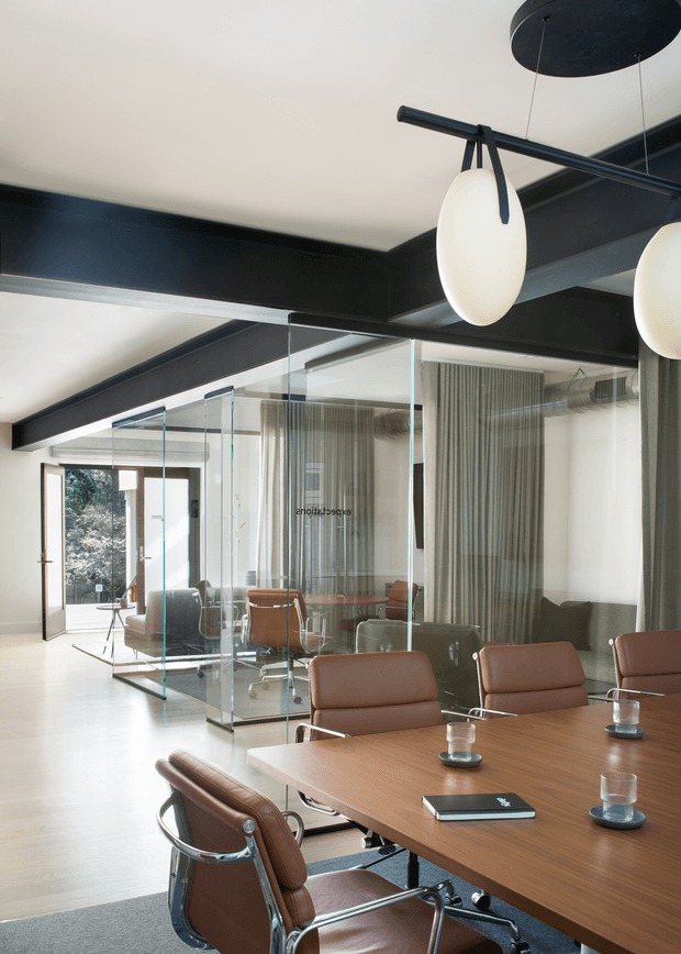Contemporary corporate meeting room with a large wood conference table and brown leather swivel chairs. The space features full-height glass partitions, exposed dark structural beams, pale timber flooring, sheer curtains, and sculptural oval pendant lights. Daylight enters through glazed doors at the rear, creating a refined, professional atmosphere.