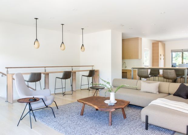 Bright open-plan contemporary living area with a large light-coloured sectional sofa, textured area rug, low timber coffee table, and mid-century-style lounge chair. A timber breakfast bar with stools separates the living zone from a compact kitchen featuring light timber cabinetry, pale benchtops, and pendant lighting. White walls and ceiling, pale flooring, and abundant daylight create an airy, refined atmosphere.