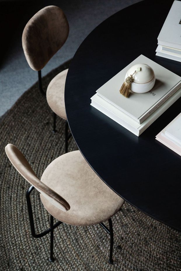 Top-down interior detail of a refined dining area, featuring a round black table with stacked neutral-toned books and a small ceramic decorative vessel, surrounded by upholstered beige dining chairs with black metal legs. A textured dark patterned carpet covers the floor, creating a sophisticated, moody setting.