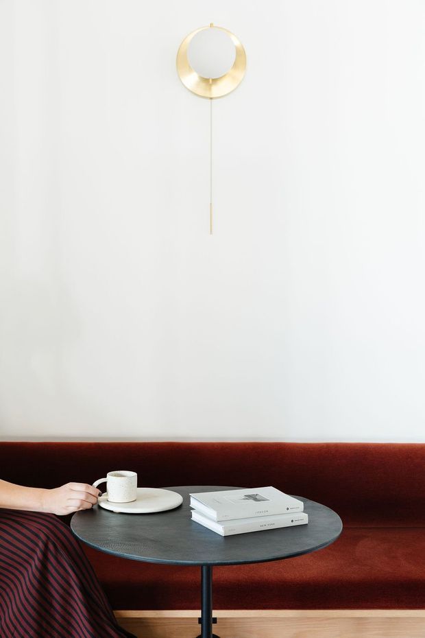 Minimalist interior vignette featuring a round dark coffee table with a white cup and saucer and stacked books, positioned in front of a deep red upholstered bench or sofa. A sculptural circular brass wall light with a slender vertical stem is mounted on the white wall above. The scene has a refined, editorial feel with strong red, white, dark grey, and brass-gold accents.