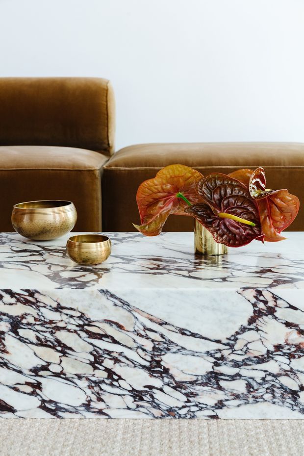 Close-up interior view of a richly veined white marble coffee table, with metallic gold-toned bowls and a floral arrangement in a reflective vessel. A brown leather sofa sits behind the table against a pale wall, creating a refined, luxurious living-room vignette.