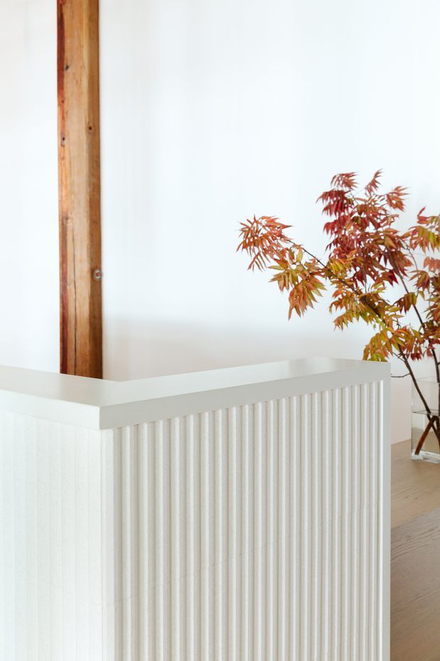 Bright minimalist interior detail showing a white vertically fluted partition or balustrade in the foreground, pale timber flooring, white walls, and a prominent exposed warm timber structural post. A reddish-leaved indoor plant adds natural colour at the right.