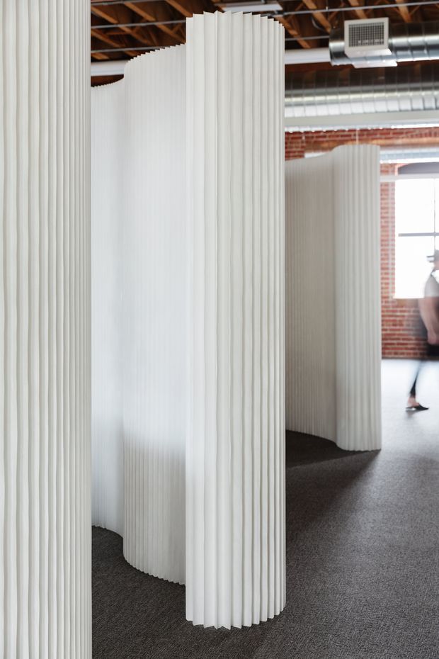 Interior view of a large open-plan commercial space featuring tall, sculptural white ribbed partition screens that curve and meander to create semi-enclosed zones. The space has dark carpet flooring, exposed timber roof framing, visible metal HVAC ductwork, and exposed red brick walls with large industrial-style windows. A person is partially visible in the background.