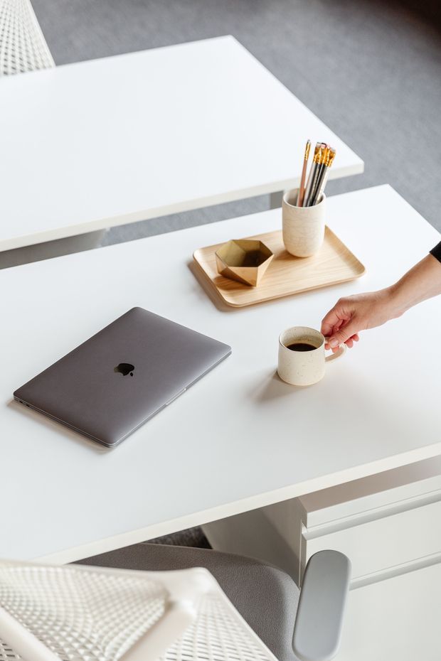 Bright, minimalist home office scene viewed from above, featuring a white desk with a closed laptop, ceramic coffee cup, wooden tray with stationery, and a modern white office chair. The surrounding surfaces are clean and uncluttered, with soft neutral tones and contemporary furnishings.