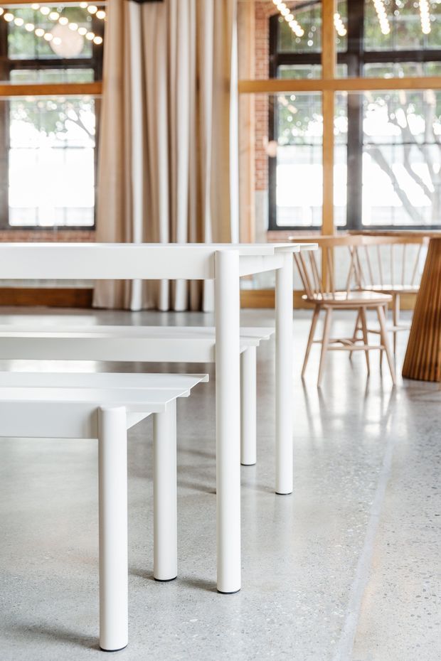 Bright interior featuring a row of simple white rectangular tables with cylindrical legs, polished light terrazzo flooring, exposed brick window surrounds, timber-framed industrial-style glazing, and warm beige curtains. Wooden spindle-back chairs and decorative string lights add warmth to the airy space.