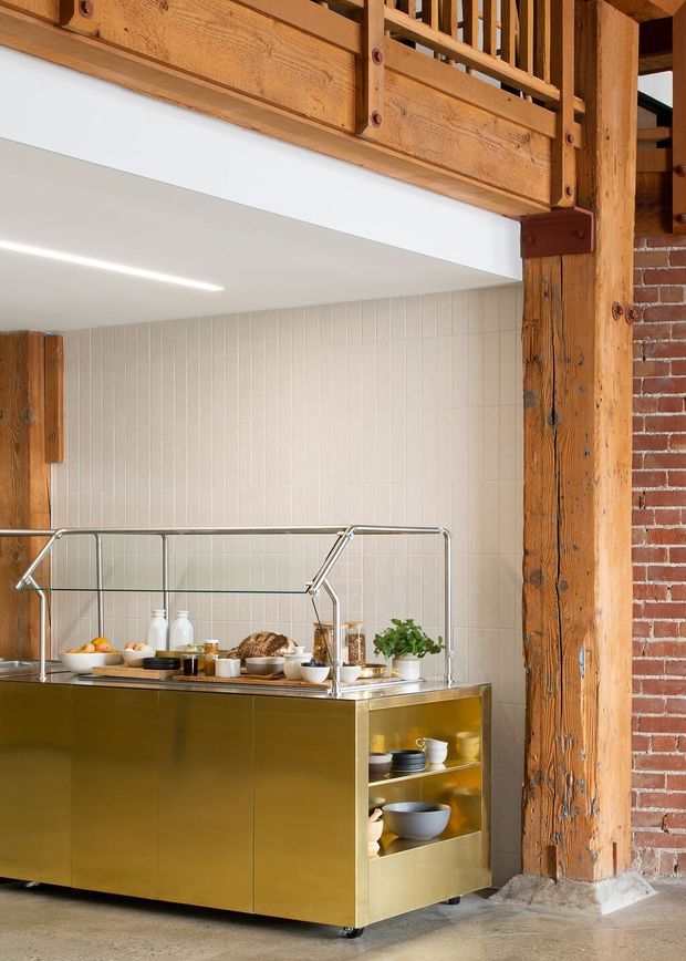 A close interior view of a commercial-style kitchen or serving area set within a converted industrial structure. A polished brass-gold island with open shelving and a stainless-steel framed glass guard displays bowls, bottles, bread and plants. Behind it is a pale vertically grooved tile wall, while exposed timber beams and columns, steel brackets, exposed red brick and a raw concrete floor reinforce the industrial character. A clean linear ceiling light provides even illumination.