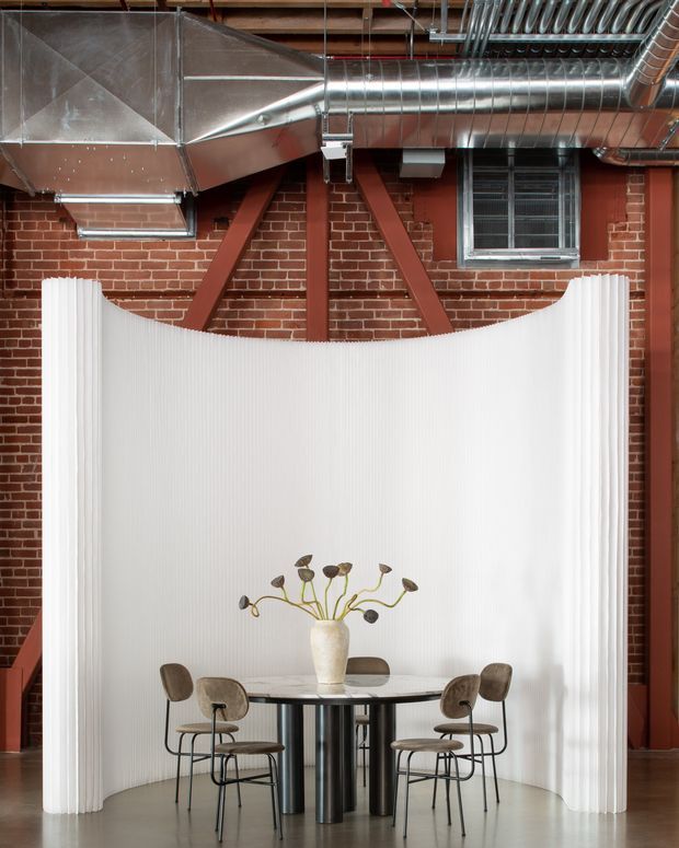 A dining area in an industrial-style interior, framed by exposed red brick walls, dark structural braces, and visible metal HVAC ductwork. A sculptural white ribbed partition curves behind a round dining table with a pale stone-like top and dark cylindrical pedestal legs, surrounded by six brown upholstered chairs. A simple vase with dried botanical stems forms the table centrepiece.