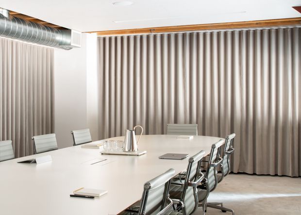 Bright contemporary conference room with a large white boardroom table surrounded by modern metal-and-leather office chairs. Full-height taupe curtains cover the walls, with exposed silver ventilation ductwork and warm timber ceiling trim adding an industrial accent. A polished metal carafe and glassware sit on a tray at the centre of the table.