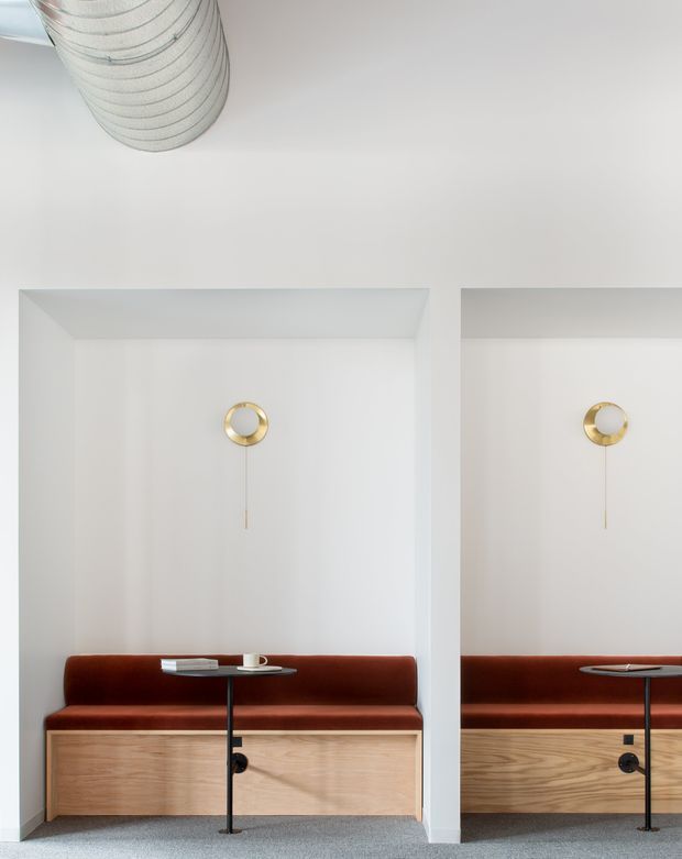 A minimalist built-in seating and work nook with a deep rust-red upholstered bench set above light timber cabinetry. White recessed wall bays frame small round brass wall lights, while slim black pedestal tables and grey carpet complete the refined, highly pared-back interior.