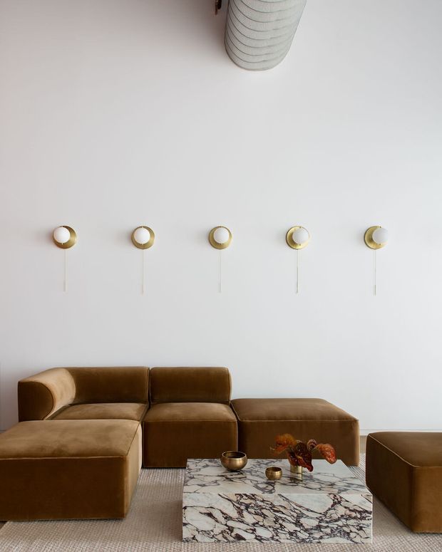 A refined living room with a low modular brown leather sectional sofa and matching ottoman seating arranged around a rectangular marble coffee table. The predominantly white wall features five evenly spaced circular brass-and-white wall lights, while a ribbed cylindrical pendant or ceiling fixture is partially visible above. The room has a restrained, luxurious palette with textured neutral carpeting and minimal decorative objects.