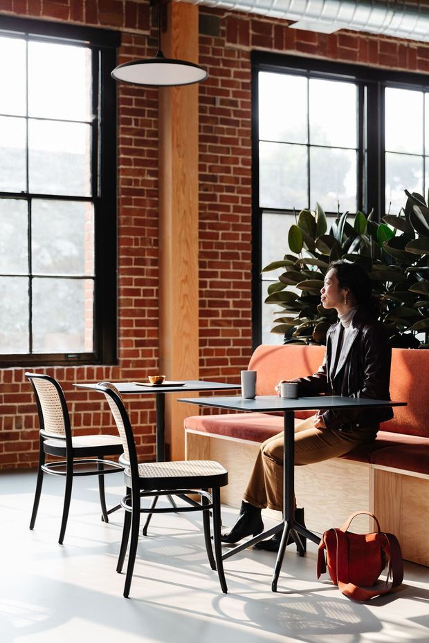 A bright industrial-style café or commercial lounge with exposed red brick walls, large black-framed multi-pane windows, exposed ductwork, timber structural columns, and built-in upholstered bench seating. Small black tables and woven-seat chairs occupy the foreground, with a person seated at the banquette and large indoor plants behind it.