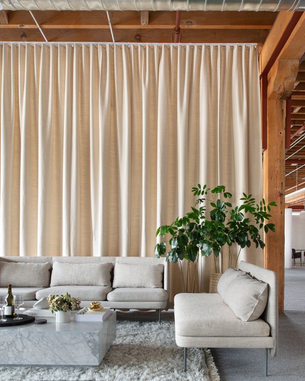 A refined open-plan living room with a large light-grey sectional sofa, matching upholstered lounge chair, marble coffee table, and soft textured rug. Full-height cream curtains span the rear wall, while exposed timber beams, timber posts, and visible metal ductwork create a warm industrial loft character. A large indoor plant adds greenery beside the seating area, and a dining or workspace zone is visible beyond the timber structural frame.