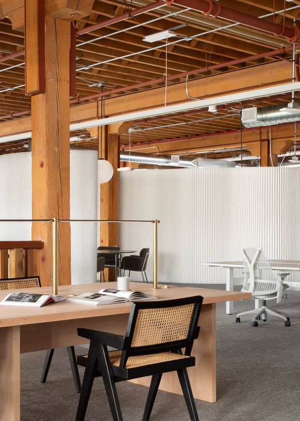 An open-plan commercial office with exposed timber beams and columns, visible red fire-suppression pipes, suspended linear lighting, exposed ductwork, and grey carpet flooring. Pale ribbed translucent partitions define work zones. The foreground features a large timber communal desk with a black-framed woven cane chair, while additional desks, task chairs, and meeting furniture occupy the background. The space combines warm timber, industrial services, and contemporary office furnishings.