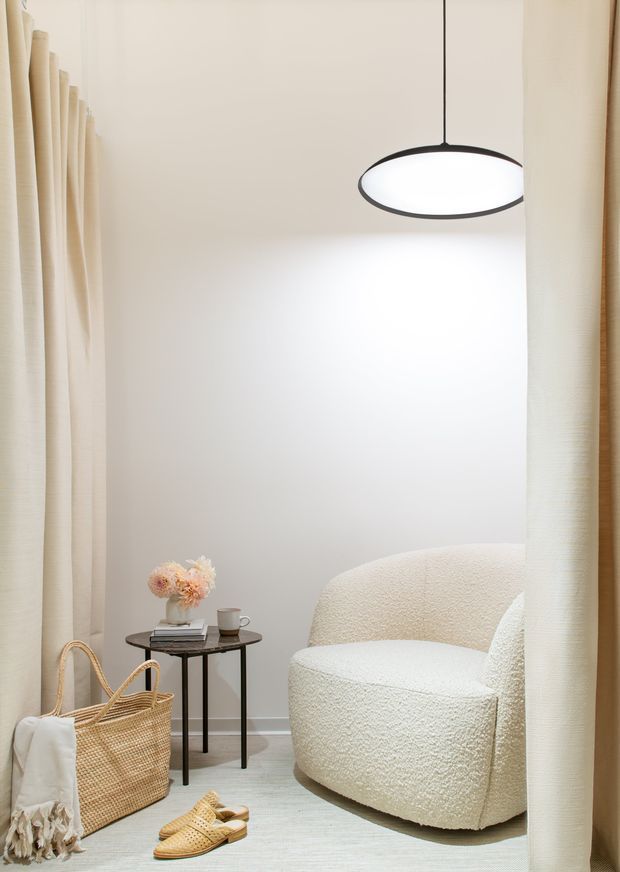A softly lit, minimalist reading nook framed by cream curtains. The space features a sculptural cream boucle armchair, a small black round side table with books, a cup and flowers, a woven basket with a light textile, and woven sandals on a pale rug. A simple oval pendant light with a black edge hangs overhead, creating a calm, warm and refined atmosphere.
