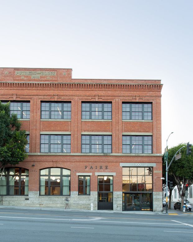 Street-facing multi-storey historic commercial building with a red brick facade, large regularly gridded industrial-style windows, decorative brick cornice and detailing, and a stone-clad lower level. Ground-floor storefront glazing and entrance face a city street, with signage reading “FAIRE” and “100.”