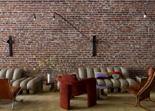 A styled living room with an exposed red brick feature wall, dramatic black articulated wall lamps, plush sculptural seating in muted neutrals and rust tones, a vintage leather sling chair, a low coffee table, and a small side table. The room has a warm, eclectic and industrial character with a low, intimate seating arrangement.