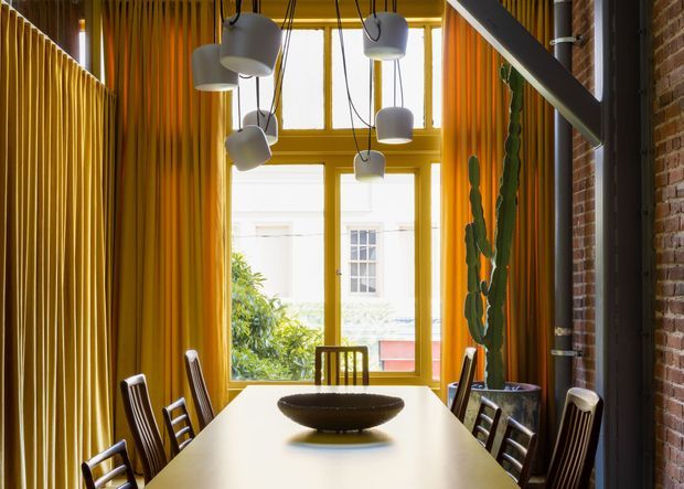 A narrow, tall dining room with a long light-coloured dining table and dark wooden chairs. Dramatic floor-to-ceiling golden curtains frame a large multi-pane window and glazed door, while clustered pendant lights hang above the table. A red brick wall, dark exposed structural beam, tall potted cactus, and a simple bowl centrepiece create a warm, industrial-contemporary atmosphere.