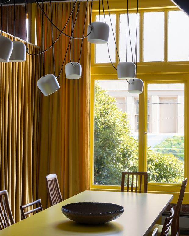 A vibrant dining room with a long pale dining table and wooden chairs beside large yellow-painted windows. Floor-to-ceiling mustard-gold curtains cover the adjacent wall, while multiple sculptural white pendant lights hang from long black cords above the table. The room has a bold, eclectic, colour-forward interior with strong natural light and views of greenery and a neighbouring building through the windows.