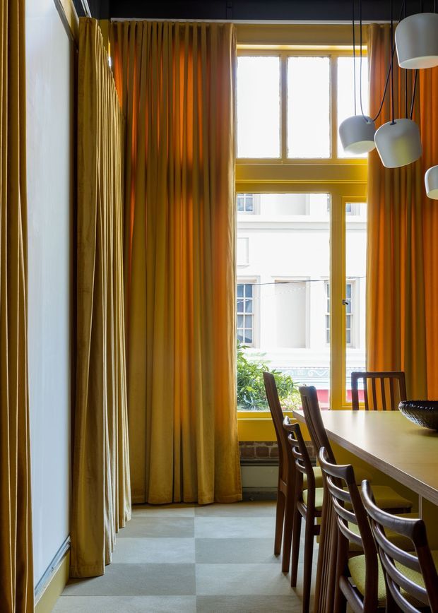 A narrow, tall dining area with a long light-coloured dining table and dark wooden chairs arranged beside expansive windows. Floor-to-ceiling golden ochre curtains frame the windows, while clustered white pendant lights hang over the table. The space has a dramatic, warm-toned palette, exposed brick at the lower wall, and large square grey floor tiles. Bright daylight enters from the tall glazed openings, with an urban building visible beyond.
