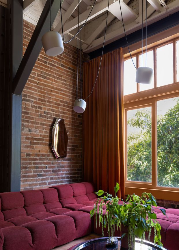 A richly styled loft-like living room with exposed red brick walls, dark structural steel framing, exposed painted timber roof beams, and a large timber-framed window. A deep burgundy modular sectional sofa wraps around a reflective coffee table with lush indoor plants. Sculptural pendant lights hang from long cords, while full-height rust-brown curtains add warmth and drama. The space combines industrial architectural elements with bold contemporary furnishings.