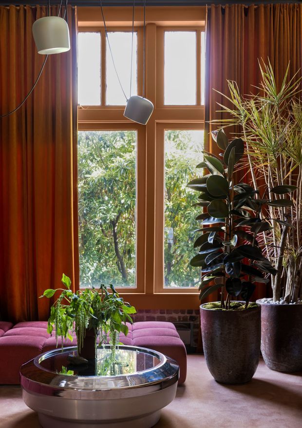 A richly styled living room with warm terracotta-toned walls and full-height drapery framing tall timber-framed windows. Two sculptural pendant lights hang in front of the glazing, while abundant large potted plants add a strong organic element. A low rounded pink upholstered sofa surrounds a reflective circular coffee table, creating a sophisticated, moody interior with layered textures and natural light.
