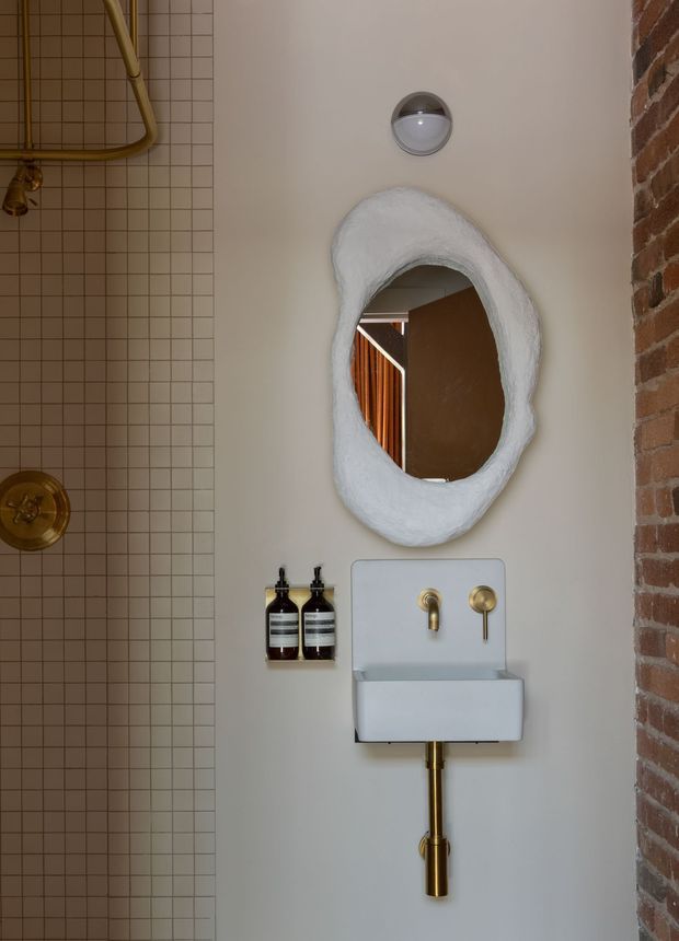 A compact bathroom with a wall-mounted white basin and exposed brass plumbing, paired with brass taps and fittings. The wall behind the basin is painted a warm off-white, with a sculptural irregular white-framed mirror above. A small square beige-tile shower zone with visible brass pipework occupies the left side, while an exposed red-brick wall forms the right edge. The space has an eclectic industrial character with warm metallic accents and a wall-mounted light.
